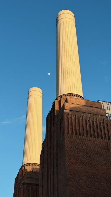 An exterior view of Battersea Power Station featuring its distinctive red brick facade and four tall, cylindrical chimneys rising against a cloudy sky. In the foreground, the River Thames is visible w
